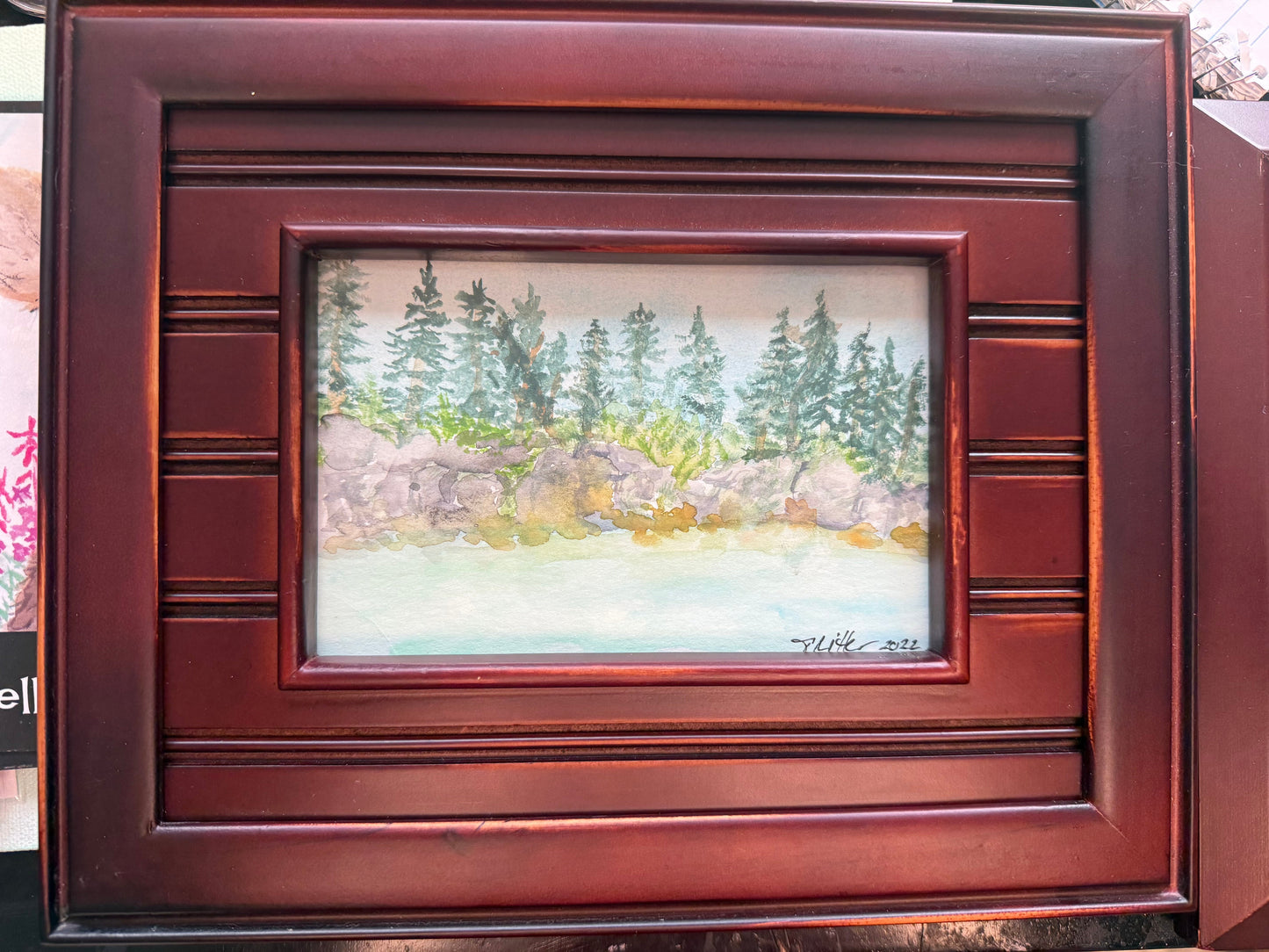 A framed landscape painting featuring green pine trees, rocks, and a pale water surface, set within a dark reddish-brown wooden frame.