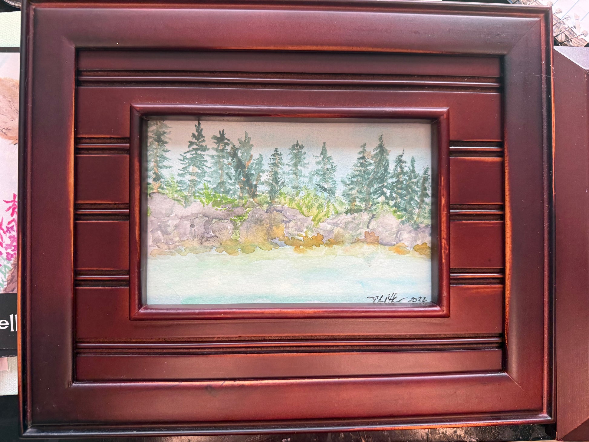A framed landscape painting featuring green pine trees, rocks, and a pale water surface, set within a dark reddish-brown wooden frame.
