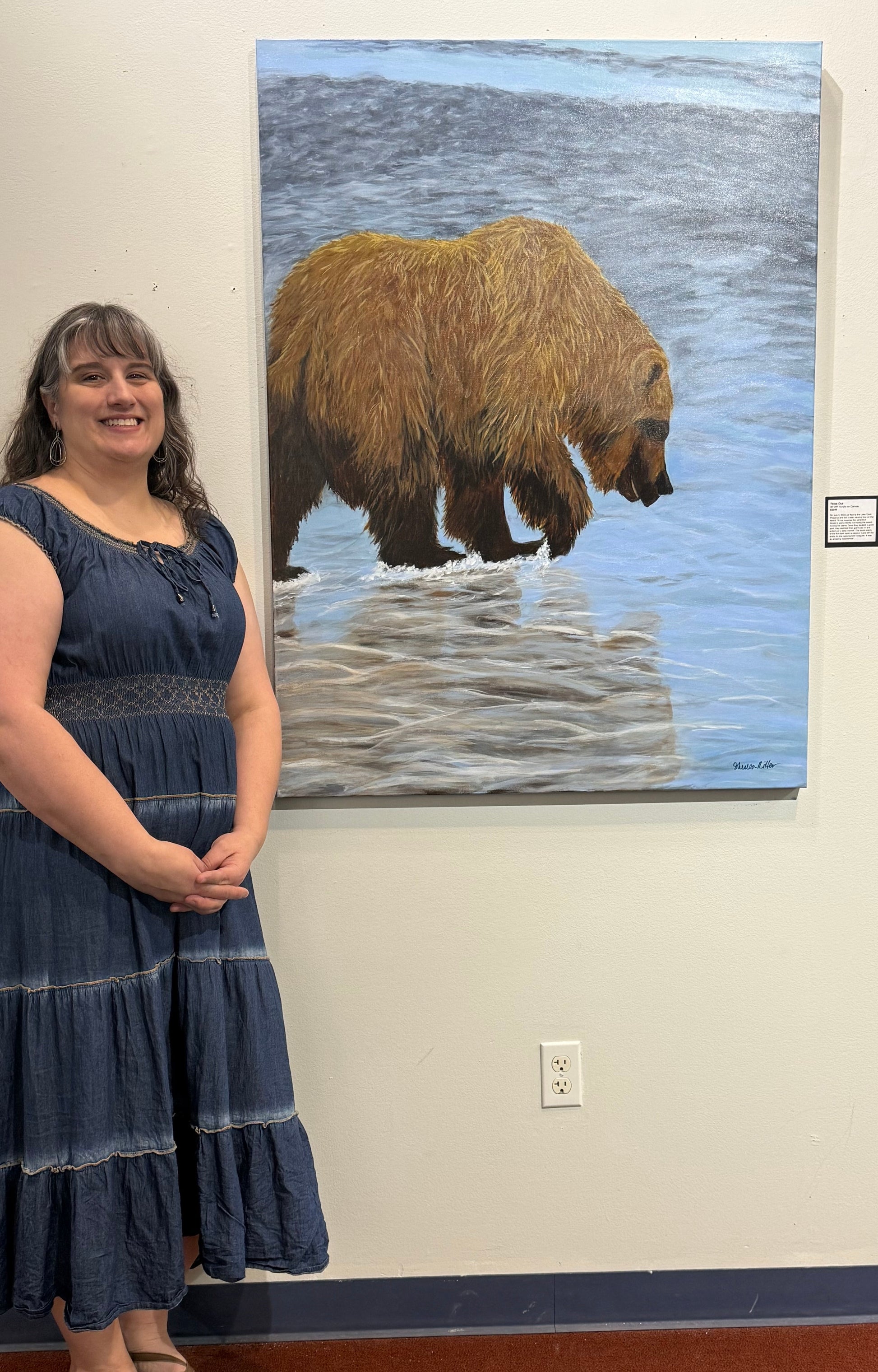 Artist posing next to her painting of large brown bear foraging for clams.